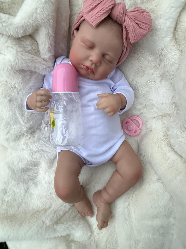 May include: A realistic baby doll with closed eyes, wearing a white long-sleeve bodysuit and a dusty pink cable-knit headband. The doll holds a clear plastic baby bottle with a pink cap and has a small pink pacifier resting nearby on the fabric.