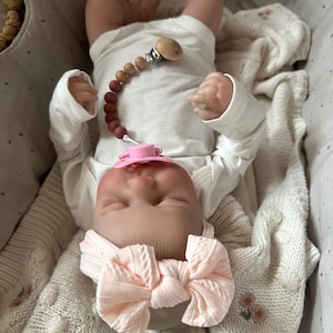 May include: A realistic baby doll wearing a long-sleeved white bodysuit and a pale pink knotted headband. The doll has a pink pacifier attached to a wooden bead pacifier clip with a gradient of brown and tan beads, resting on a cream-colored blanket.
