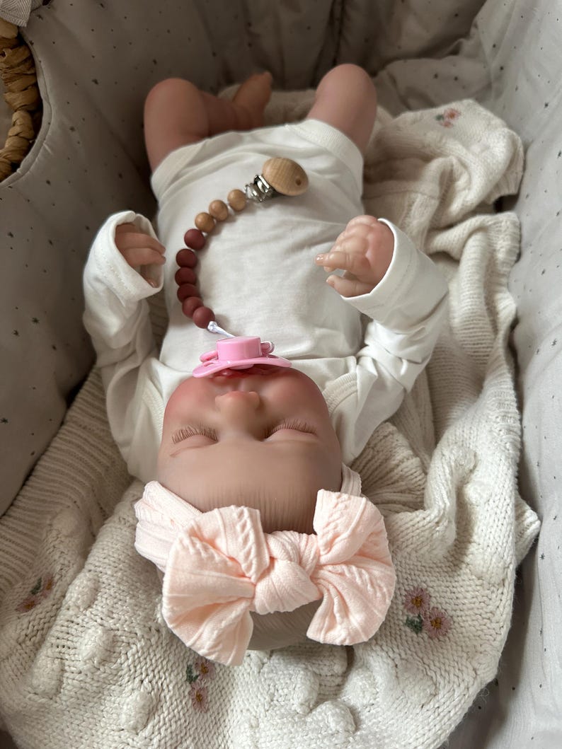 May include: A realistic baby doll wearing a long-sleeved white bodysuit and a pale pink knotted headband. The doll has a pink pacifier attached to a wooden bead pacifier clip with a gradient of brown and tan beads, resting on a cream-colored blanket.