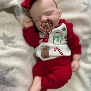 Pode incluir: Uma boneca beb&eacute; a dormir com uma fita de la&ccedil;o vermelho e uma chupeta em ouro rosa. A boneca veste uma camisola de malha vermelha e branca com padr&otilde;es de renas, bengalas doces e presentes, com cal&ccedil;as de malha vermelhas e dois bot&otilde;es.