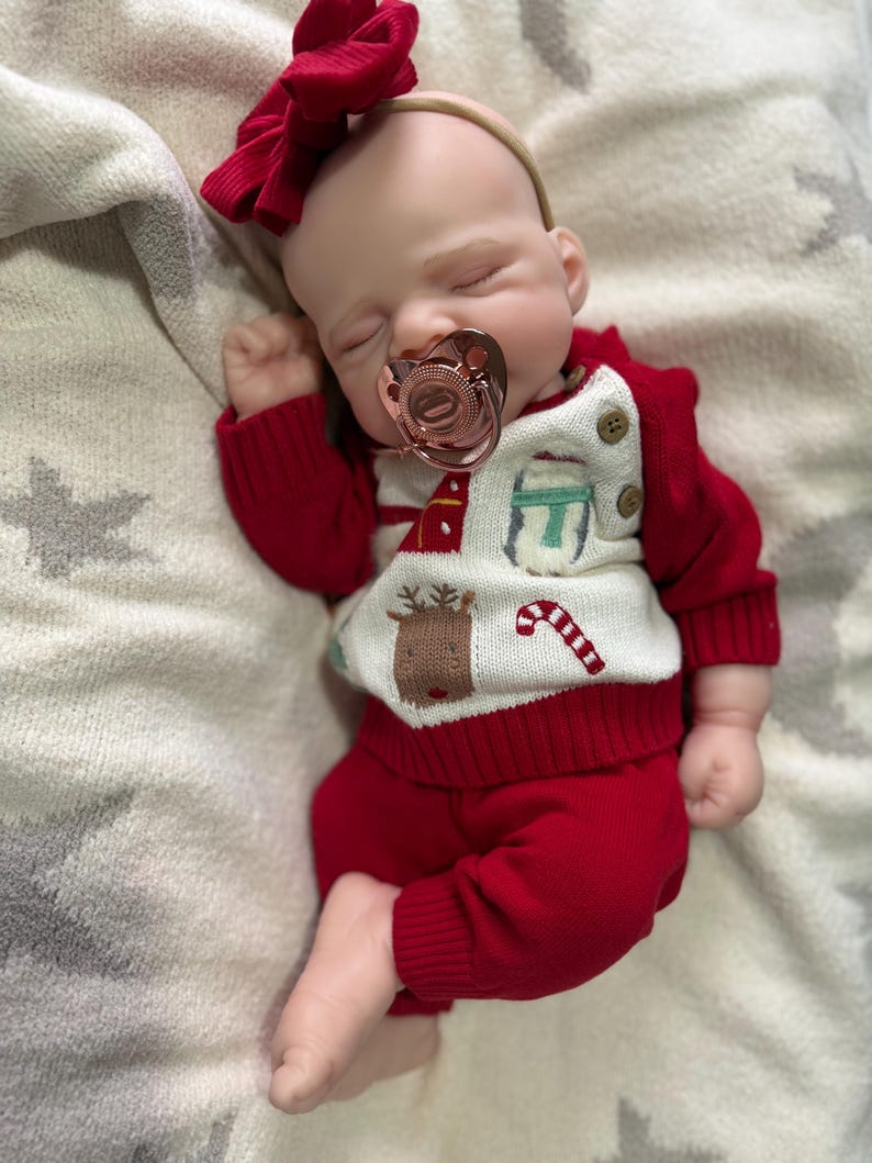 Pode incluir: Uma boneca beb&eacute; a dormir com uma fita de la&ccedil;o vermelho e uma chupeta em ouro rosa. A boneca veste uma camisola de malha vermelha e branca com padr&otilde;es de renas, bengalas doces e presentes, com cal&ccedil;as de malha vermelhas e dois bot&otilde;es.