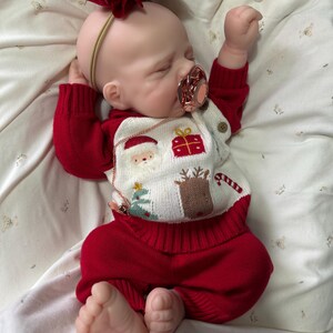 Pode incluir: Uma boneca rec&eacute;m-nascida realista com olhos fechados, vestindo um su&eacute;ter de malha vermelho e branco com motivos natalinos, incluindo Papai Noel, um presente, uma rena e uma &aacute;rvore. A boneca tem uma faixa com la&ccedil;o de veludo vermelho, cal&ccedil;as vermelhas e uma chupeta.
