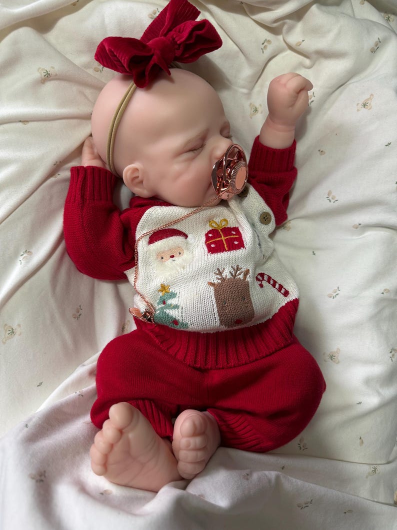 Pode incluir: Uma boneca rec&eacute;m-nascida realista com olhos fechados, vestindo um su&eacute;ter de malha vermelho e branco com motivos natalinos, incluindo Papai Noel, um presente, uma rena e uma &aacute;rvore. A boneca tem uma faixa com la&ccedil;o de veludo vermelho, cal&ccedil;as vermelhas e uma chupeta.