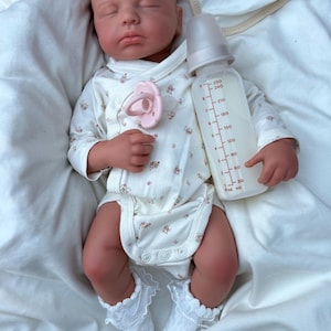 May include: A realistic baby doll dressed in a white floral onesie, white lace socks, and a pink dummy. The doll holds a clear baby bottle with milk, marked with measurements in millilitres.