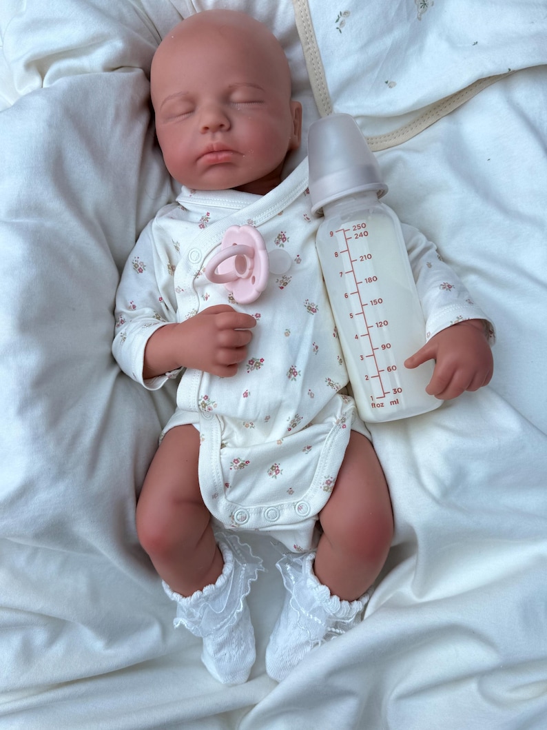 May include: A realistic baby doll dressed in a white floral onesie, white lace socks, and a pink dummy. The doll holds a clear baby bottle with milk, marked with measurements in millilitres.