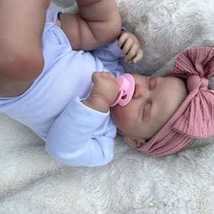 May include: A realistic baby doll with light skin, wearing a plain white long-sleeved onesie and a dusty rose pink ribbed headband featuring a large bow. The doll has closed eyes and is holding a light pink pacifier in its mouth while lying down.