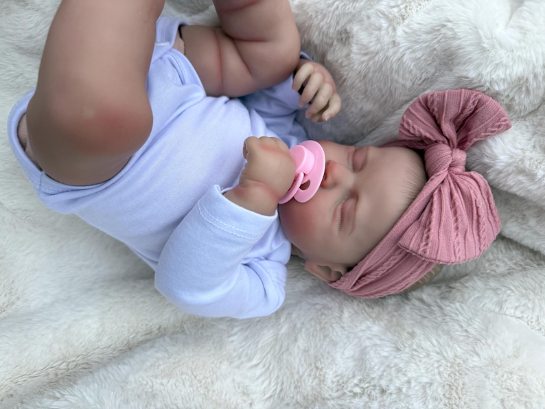 May include: A realistic baby doll with light skin, wearing a plain white long-sleeved onesie and a dusty rose pink ribbed headband featuring a large bow. The doll has closed eyes and is holding a light pink pacifier in its mouth while lying down.