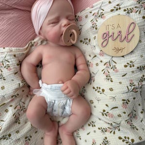 May include: A realistic newborn doll with closed eyes, wearing a light pink knotted headband and a tan pacifier. The doll is dressed in a white nappy with a grey pattern. Beside the doll is a round wooden plaque with the text It's a girl.
