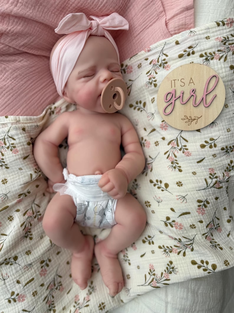 May include: A realistic newborn doll with closed eyes, wearing a light pink knotted headband and a tan pacifier. The doll is dressed in a white nappy with a grey pattern. Beside the doll is a round wooden plaque with the text It's a girl.