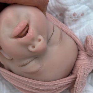 Może przedstawiać: Zbliżenie twarzy lalki dziecka z zamkniętymi oczami i lekko otwartymi ustami. Lalka ma r&oacute;żowy odcień sk&oacute;ry i nosi r&oacute;żową dzianinową czapkę. Twarz lalki jest trzymana przez dłoń.