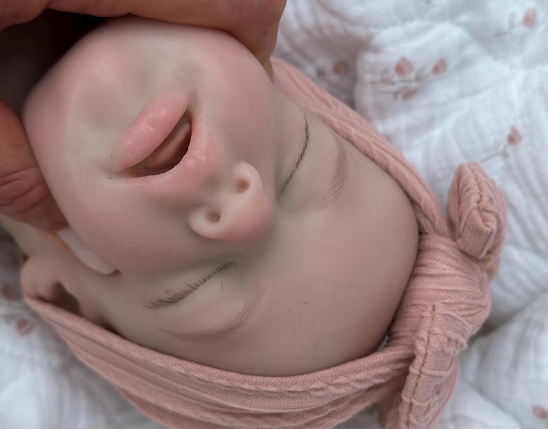 Może przedstawiać: Zbliżenie twarzy lalki dziecka z zamkniętymi oczami i lekko otwartymi ustami. Lalka ma r&oacute;żowy odcień sk&oacute;ry i nosi r&oacute;żową dzianinową czapkę. Twarz lalki jest trzymana przez dłoń.