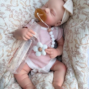 May include: A baby doll wearing a pink and white striped onesie and a cream-colored bow headband. The doll has a pacifier and a bottle. The doll is lying on a patterned blanket with a floral and animal design.