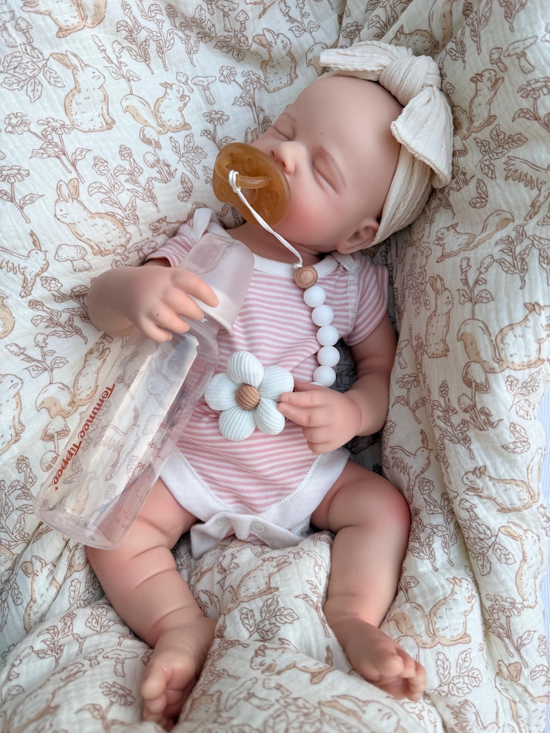 May include: A baby doll wearing a pink and white striped onesie and a cream-colored bow headband. The doll has a pacifier and a bottle. The doll is lying on a patterned blanket with a floral and animal design.