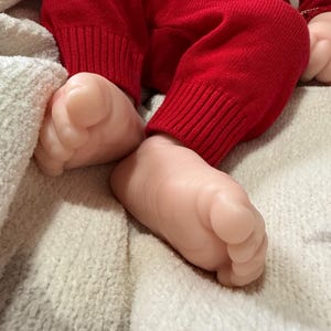 Pode incluir: Um par de p&eacute;s de boneca realistas com tons de pele claros, posicionados perto de cal&ccedil;as de malha vermelhas brilhantes com punhos canelados. A boneca repousa sobre uma superf&iacute;cie de tecido texturizado branco, mostrando dedos detalhados e pele macia.