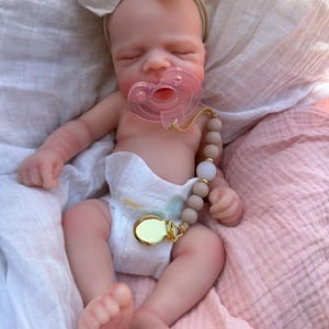 May include: A realistic newborn doll with closed eyes, wearing a beige fabric bow headband and a white diaper. The doll has a translucent pink pacifier in its mouth, attached to a beige and white beaded pacifier clip with a gold metal clasp.