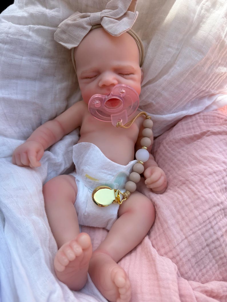May include: A realistic newborn doll with closed eyes, wearing a beige fabric bow headband and a white diaper. The doll has a translucent pink pacifier in its mouth, attached to a beige and white beaded pacifier clip with a gold metal clasp.