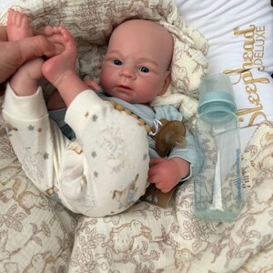 May include: A realistic baby doll with blue eyes, wearing a light blue onesie and patterned pants. A clear baby bottle and a pacifier are next to the doll. The doll is lying on a cream-colored blanket with a bunny pattern.