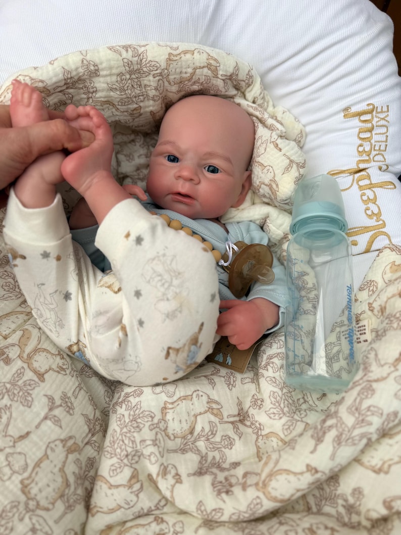 May include: A realistic baby doll with blue eyes, wearing a light blue onesie and patterned pants. A clear baby bottle and a pacifier are next to the doll. The doll is lying on a cream-colored blanket with a bunny pattern.