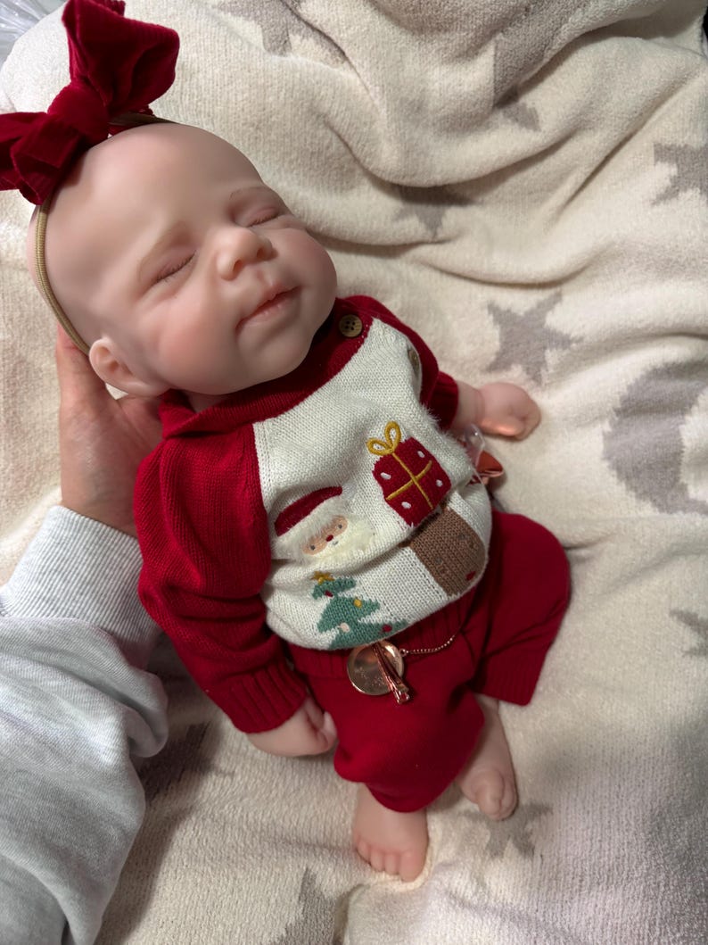 Pode incluir: Uma boneca realista com olhos fechados, usando uma faixa com la&ccedil;o de veludo vermelho e um su&eacute;ter de tric&ocirc; creme com um gr&aacute;fico de Papai Noel, uma &aacute;rvore de Natal e um presente, combinada com cal&ccedil;as de tric&ocirc; vermelhas e um pingente met&aacute;lico.
