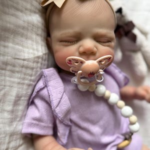 Pu&ograve; includere: Una bambola neonata realistica con gli occhi chiusi, che indossa una maglietta a maniche corte color viola chiaro con spalle arricciate e una fascia beige con fiocco. La bambola ha un ciuccio rosa chiaro con un disegno di fragola e una catenella portaciuccio con perline bianche e beige.