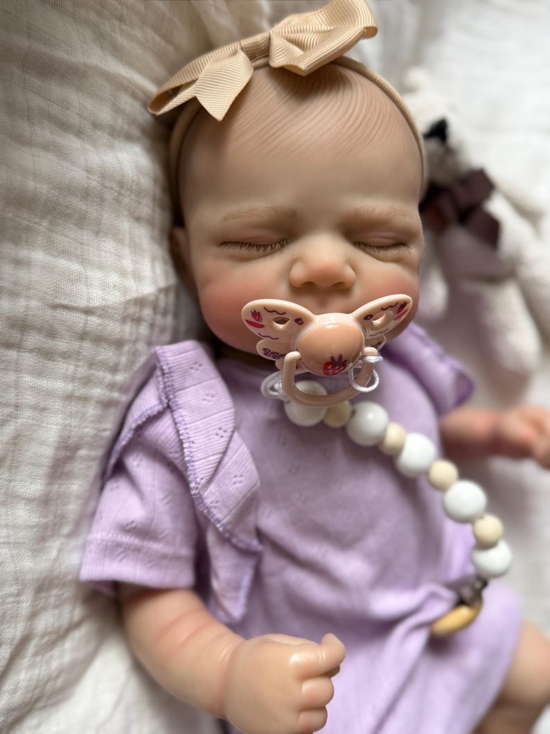 Pu&ograve; includere: Una bambola neonata realistica con gli occhi chiusi, che indossa una maglietta a maniche corte color viola chiaro con spalle arricciate e una fascia beige con fiocco. La bambola ha un ciuccio rosa chiaro con un disegno di fragola e una catenella portaciuccio con perline bianche e beige.