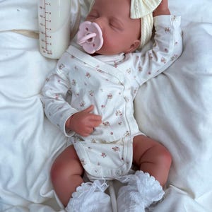 May include: A realistic baby doll dressed in a white floral onesie, a matching headband with a bow, and lace-trimmed socks. The doll has a pink dummy and lies next to a bottle filled with milk. The doll's skin has a realistic tone.
