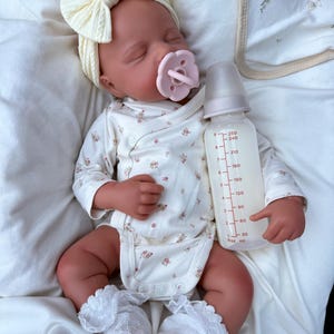 May include: A sleeping baby doll wearing a cream bow headband, a floral print onesie, and white lace-trimmed socks. A pink dummy is in its mouth, and a baby bottle with liquid is held nearby.