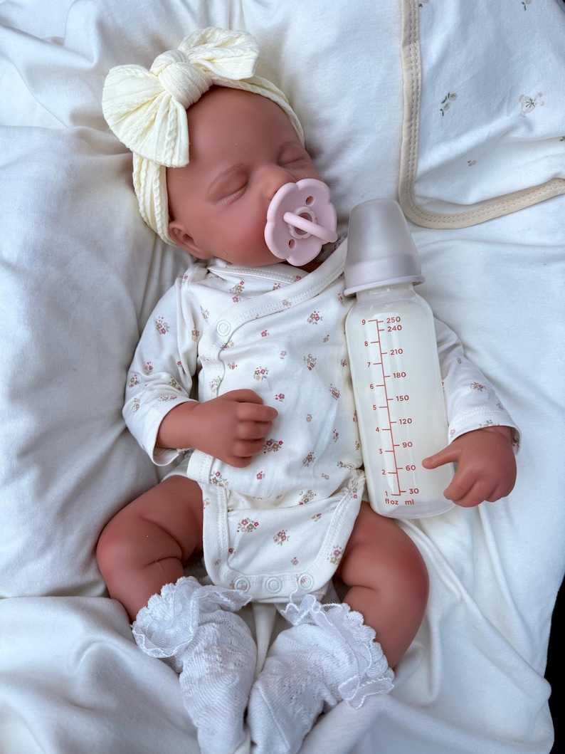 May include: A sleeping baby doll wearing a cream bow headband, a floral print onesie, and white lace-trimmed socks. A pink dummy is in its mouth, and a baby bottle with liquid is held nearby.
