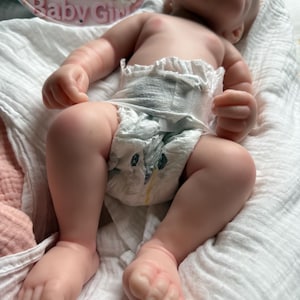 Pu&ograve; includere: Una bambola realistica dalla pelle chiara, sdraiata su un panno di mussola bianco. La bambola indossa un pannolino usa e getta con un piccolo disegno di pinguino. Sullo sfondo, un cartello circolare presenta fiori rosa e il testo Welcome Baby Girl.