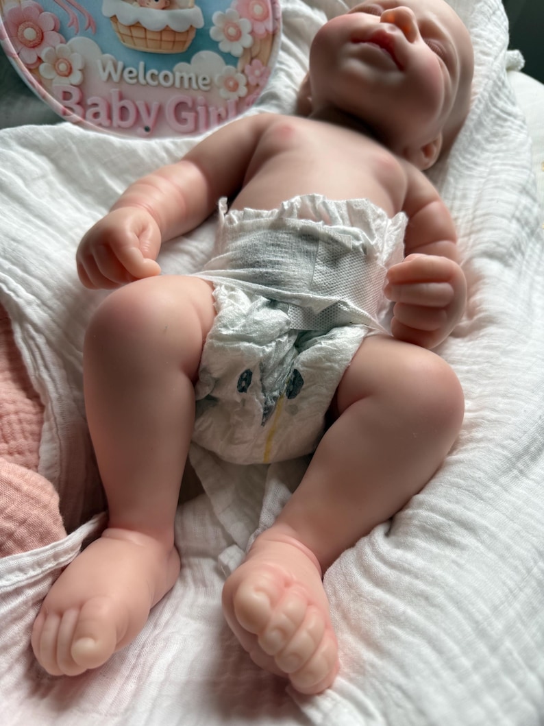 Pu&ograve; includere: Una bambola realistica dalla pelle chiara, sdraiata su un panno di mussola bianco. La bambola indossa un pannolino usa e getta con un piccolo disegno di pinguino. Sullo sfondo, un cartello circolare presenta fiori rosa e il testo Welcome Baby Girl.