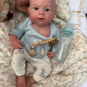 May include: A realistic baby doll with a bald head, blue eyes, and rosy cheeks. The doll is wearing a light blue onesie with a bunny graphic and matching pants. A pacifier clip and a baby bottle are also visible. The doll is resting on a patterned blanket.