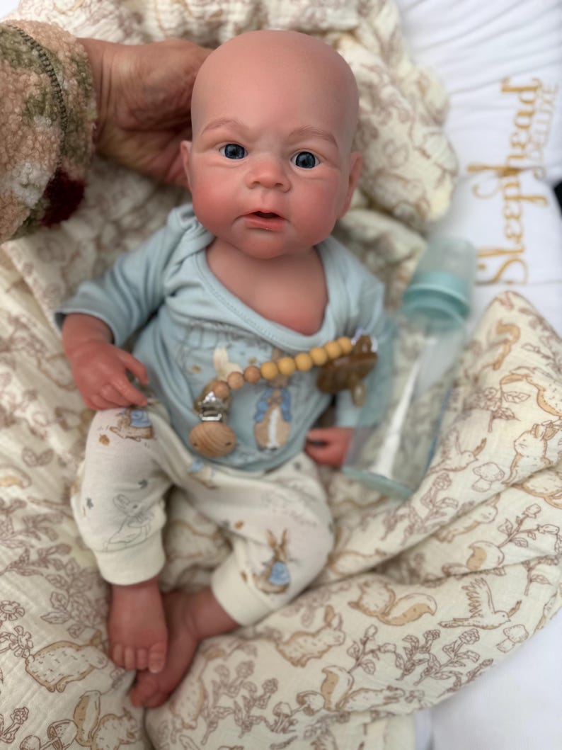 May include: A realistic baby doll with a bald head, blue eyes, and rosy cheeks. The doll is wearing a light blue onesie with a bunny graphic and matching pants. A pacifier clip and a baby bottle are also visible. The doll is resting on a patterned blanket.