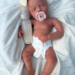 May include: A realistic baby doll with a dummy and a bottle of milk. The doll is wearing a nappy and a cream-coloured headband with a bow. The baby is lying on a white blanket with a floral pattern.
