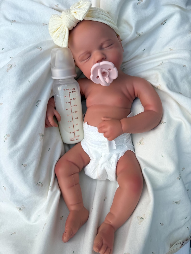 May include: A realistic baby doll with a dummy and a bottle of milk. The doll is wearing a nappy and a cream-coloured headband with a bow. The baby is lying on a white blanket with a floral pattern.