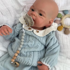 K&ouml;nnte beinhalten: Eine realistische Babypuppe mit hellem Teint, bekleidet mit einem hellblauen Zopfmuster-Pullover und passender Hose. Die Puppe tr&auml;gt einen wei&szlig;en Kragen mit kleiner brauner B&auml;renstickerei, einen beigen Schnuller und eine Schnullerkette aus grauen Holzperlen.