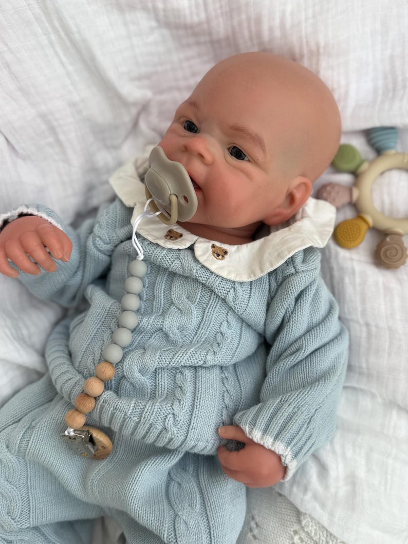K&ouml;nnte beinhalten: Eine realistische Babypuppe mit hellem Teint, bekleidet mit einem hellblauen Zopfmuster-Pullover und passender Hose. Die Puppe tr&auml;gt einen wei&szlig;en Kragen mit kleiner brauner B&auml;renstickerei, einen beigen Schnuller und eine Schnullerkette aus grauen Holzperlen.