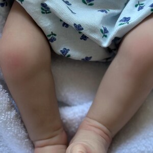 May include: A pair of realistic doll legs with light skin tone, featuring detailed toes and ankles. The legs are dressed in a white fabric garment adorned with a repeating pattern of small blue flowers and green leaves, resting on a soft white texture.
