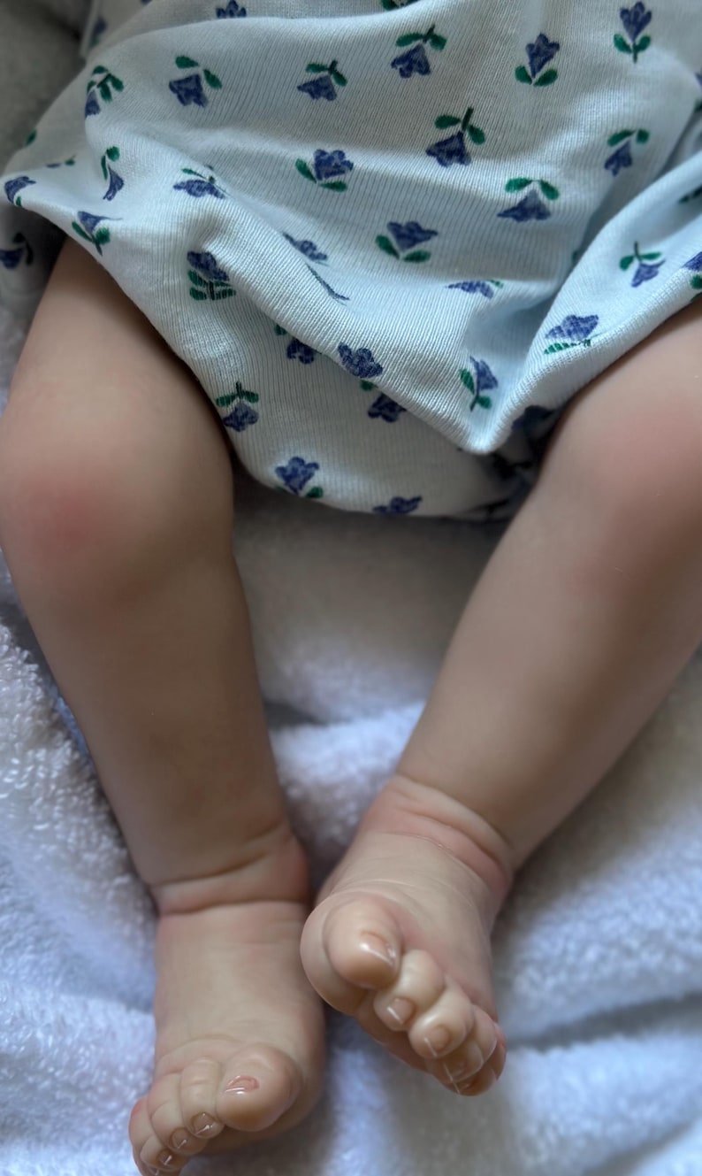 May include: A pair of realistic doll legs with light skin tone, featuring detailed toes and ankles. The legs are dressed in a white fabric garment adorned with a repeating pattern of small blue flowers and green leaves, resting on a soft white texture.