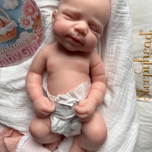 Pu&ograve; includere: Una bambola neonato realistica con pelle chiara, occhi chiusi e un sorriso accennato, che indossa un pannolino usa e getta bianco. La bambola &egrave; posizionata su un tessuto bianco accanto a un cartoncino circolare con fiori rosa e il testo Welcome Baby Girl.