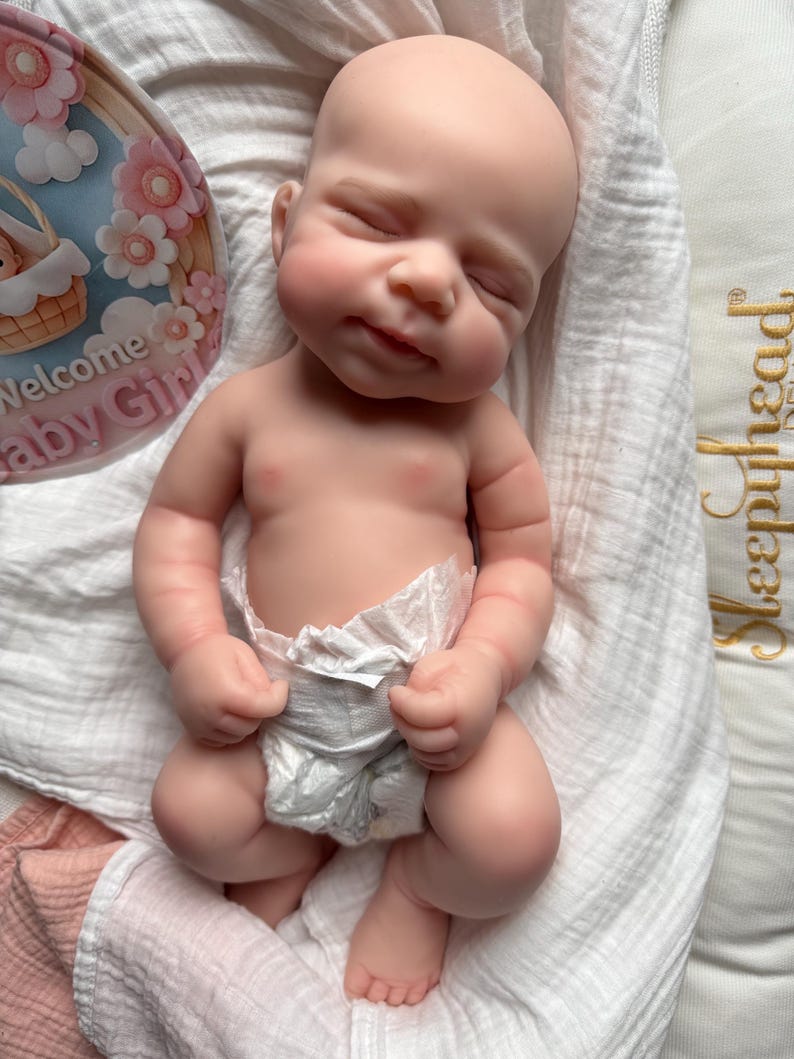 Pu&ograve; includere: Una bambola neonato realistica con pelle chiara, occhi chiusi e un sorriso accennato, che indossa un pannolino usa e getta bianco. La bambola &egrave; posizionata su un tessuto bianco accanto a un cartoncino circolare con fiori rosa e il testo Welcome Baby Girl.