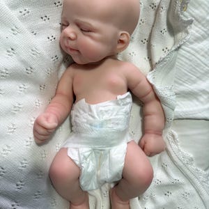 Pode incluir: Uma boneca realista de rec&eacute;m-nascido com pele clara e lisa, olhos fechados e uma express&atilde;o facial suave. A boneca &eacute; careca e usa uma fralda descart&aacute;vel branca. A figura est&aacute; posicionada deitada de costas com bra&ccedil;os e pernas dobrados.