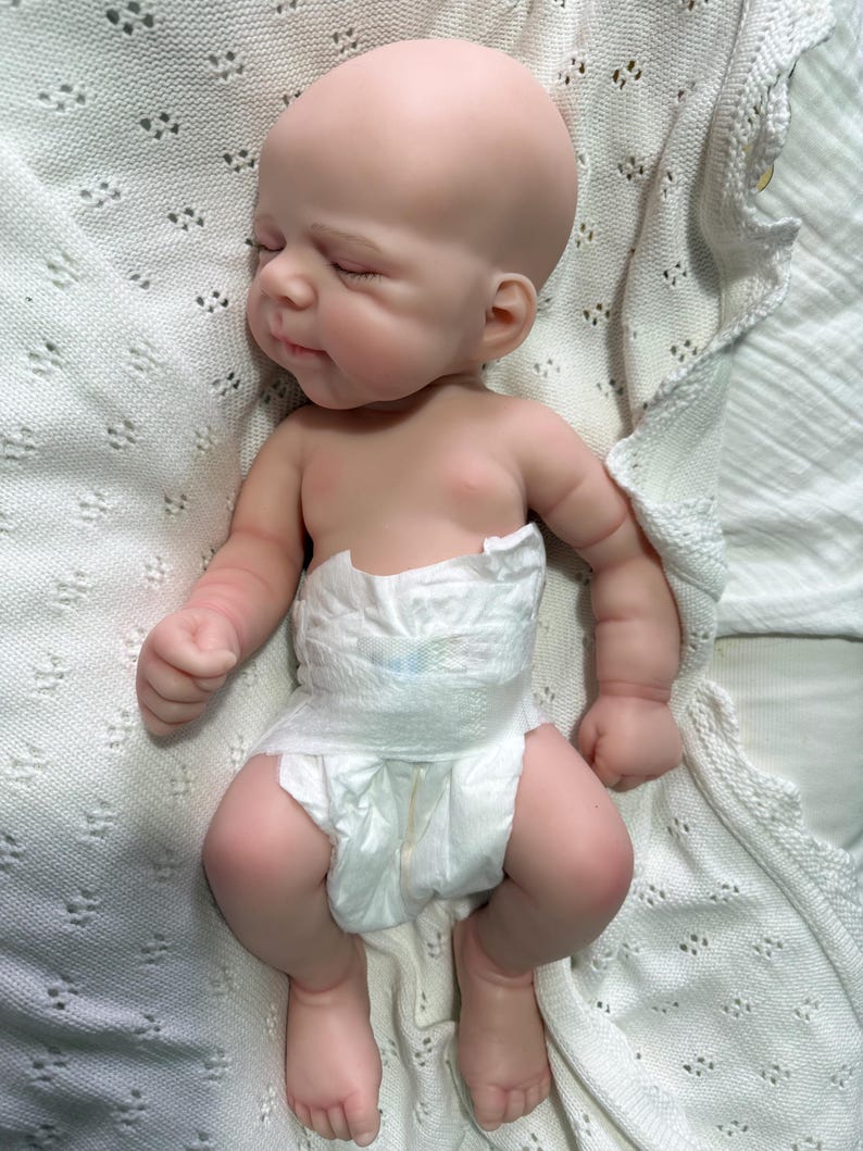 Pode incluir: Uma boneca realista de rec&eacute;m-nascido com pele clara e lisa, olhos fechados e uma express&atilde;o facial suave. A boneca &eacute; careca e usa uma fralda descart&aacute;vel branca. A figura est&aacute; posicionada deitada de costas com bra&ccedil;os e pernas dobrados.