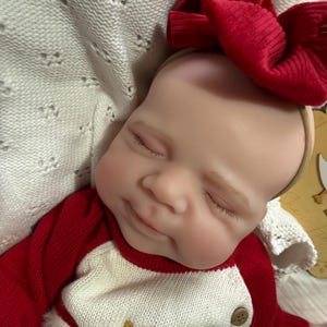 Pode incluir: Uma boneca realista com olhos fechados e um sorriso subtil, vestindo uma camisola de malha de manga comprida vermelha e branca e uma fita de cabelo grande com la&ccedil;o vermelho canelado. A boneca tem pele clara e est&aacute; sobre tecido branco.