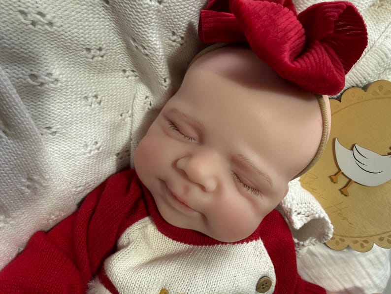 Pode incluir: Uma boneca realista com olhos fechados e um sorriso subtil, vestindo uma camisola de malha de manga comprida vermelha e branca e uma fita de cabelo grande com la&ccedil;o vermelho canelado. A boneca tem pele clara e est&aacute; sobre tecido branco.