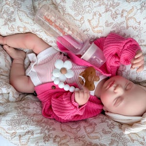 May include: A sleeping baby doll wearing a pink sweater, striped shirt, and a white headband. A clear bottle with a pink nipple is placed near the doll. A pacifier and a flower-shaped teether are also visible. The doll is lying on a patterned blanket.