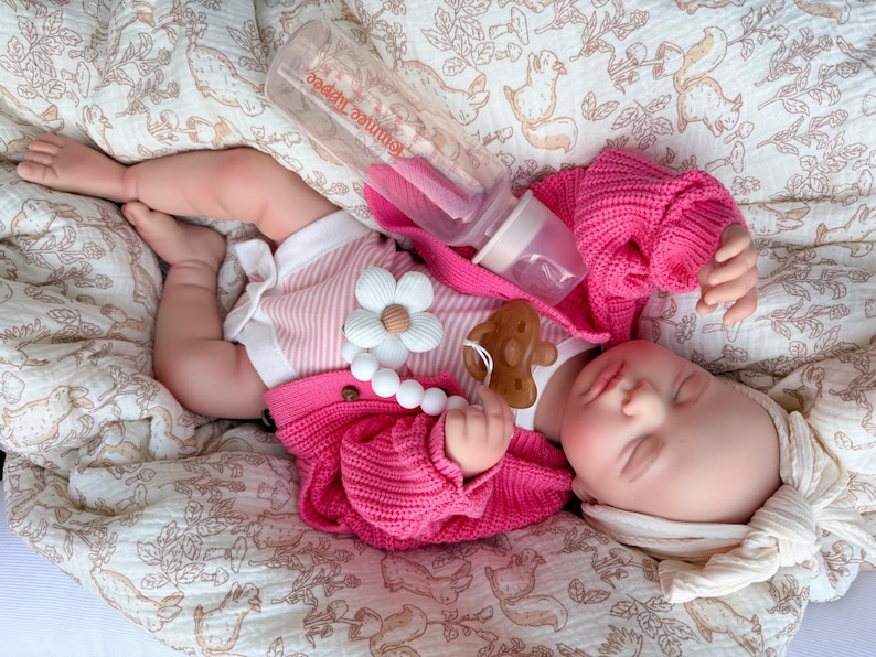 May include: A sleeping baby doll wearing a pink sweater, striped shirt, and a white headband. A clear bottle with a pink nipple is placed near the doll. A pacifier and a flower-shaped teether are also visible. The doll is lying on a patterned blanket.
