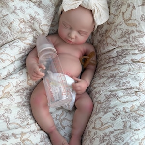 May include: A lifelike baby doll with a cream-colored bow headband, lying on a patterned blanket. The doll holds a clear bottle and has a pacifier. The blanket features a repeating pattern of squirrels and foliage.