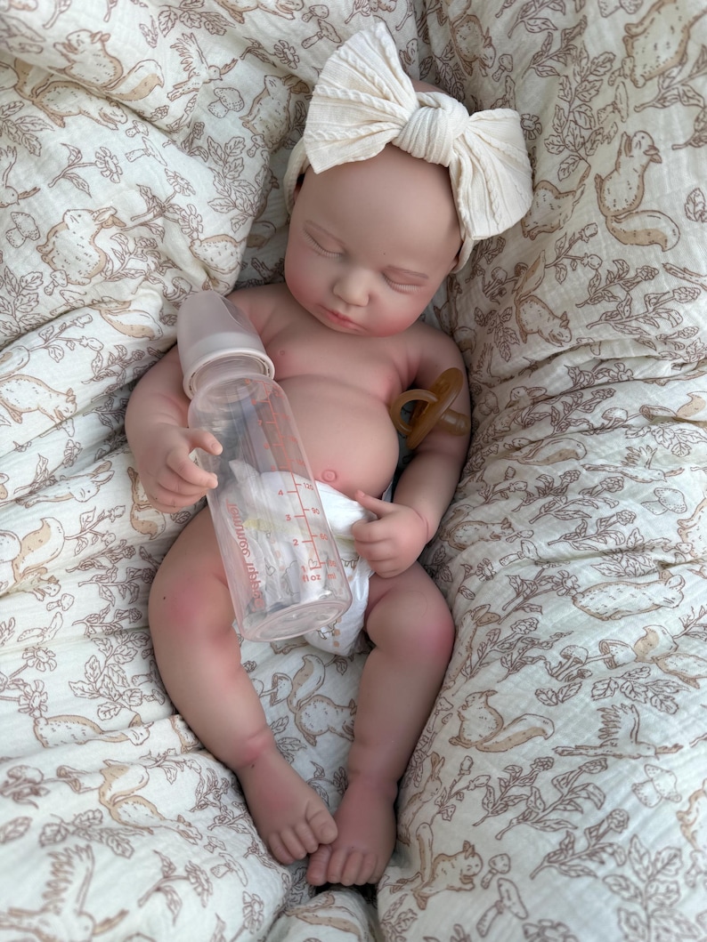 May include: A lifelike baby doll with a cream-colored bow headband, lying on a patterned blanket. The doll holds a clear bottle and has a pacifier. The blanket features a repeating pattern of squirrels and foliage.