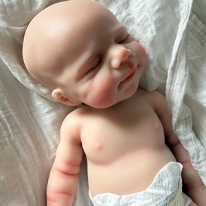 May include: A realistic reborn baby doll with pale skin, closed eyes, and a subtle smile. The doll has a smooth, hairless head, defined facial features, and soft pink cheeks. It is wearing a white disposable diaper and is wrapped in a white cloth.