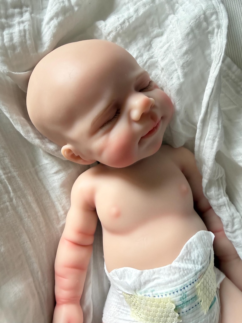 May include: A realistic reborn baby doll with pale skin, closed eyes, and a subtle smile. The doll has a smooth, hairless head, defined facial features, and soft pink cheeks. It is wearing a white disposable diaper and is wrapped in a white cloth.